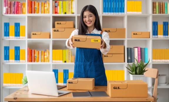 Woman handing out a GLS parcel. She stands at a desk in front of shelves field with colourful books.